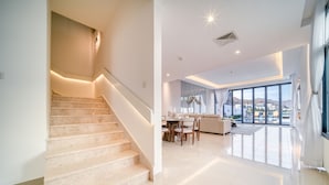 A spacious living area with a staircase leading to a balcony, large windows offering a view, and a dining area with a table and chairs.