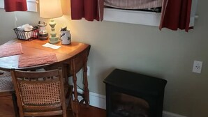 A dining area with a wooden table, a wicker chair, a small black stool, a lamp, and a window with striped curtains.