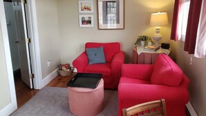 A living room with two red sofas, a small round ottoman, a wicker chair, a side table with a lamp, and framed pictures on the wall.