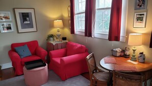 A living room with two red sofas, a wooden dining table with chairs, a small side table with a lamp, and a window with curtains.
