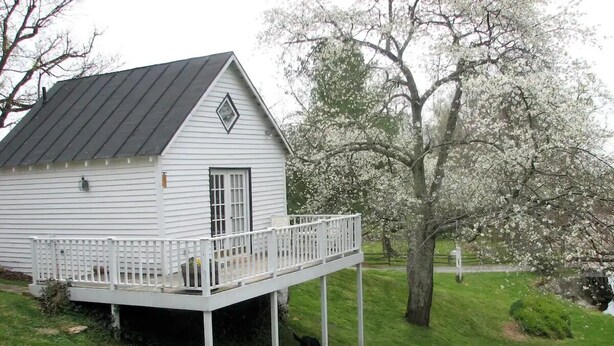 A white wooden house with a balcony, surrounded by a grassy area and blooming trees.