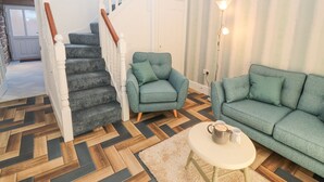 A living room with a herringbone floor pattern, a staircase with white railings, a green sofa, a matching armchair, and a small round table with a mug. - Hillside Hideaway (Wigton)