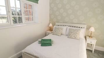 A neatly made bed with white bedding and green towels, two bedside tables with lamps, a window with a green valance, and patterned wallpaper.