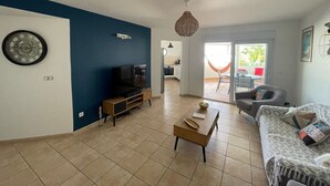 Una sala de estar con televisor, una mesa de centro y un sofá. Se ve una cocina al fondo.