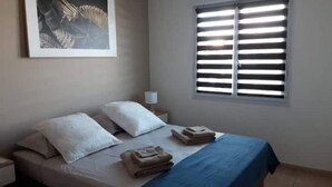 Una cama bien hecha con almohadas blancas y toallas dobladas, una obra de arte enmarcada en la pared y una ventana con persianas horizontales.