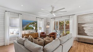 Un salon avec un grand canapé, un ventilateur de plafond et une vue sur la plage par les fenêtres.