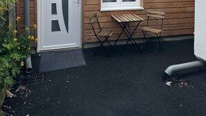 Eine Holzwand mit einer weißen Tür, einem mit Vorhängen versehenen Fenster und einer kleinen Außenbereich-Sitzgelegenheit mit zwei Holzstühlen und einem Tisch.
