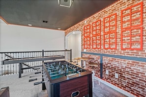 A room with a foosball table, a brick wall with red-framed pictures, and a staircase with a railing. - 5BR Home w/ Game & Themed Rooms at Champions Gate (Kissimmee)