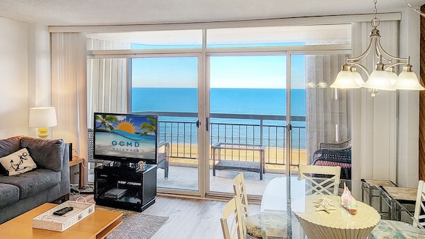 Una sala de estar con un televisor mostrando 'OCMD GETAWAYS', un sofá, una mesa de centro y un comedor con sillas y una mesa de vidrio.