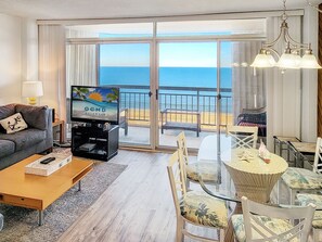 A living room with a television displaying a beach scene, a sofa, a coffee table, and a dining area with chairs and a glass table. - Oceanfront Condo steps to the beach! Pool, tennis courts, pickleball courts and more! Fountainhead Towers #1109 (Ocean City)