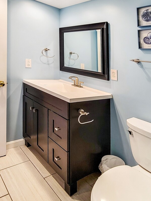 A bathroom with a dark wooden vanity, a white sink, and a mirror above it. There are two towel rings on the wall and a toilet visible. - Oceanfront Condo steps to the beach! Pool, tennis courts, pickleball courts and more! Fountainhead Towers #1109 (Ocean City)