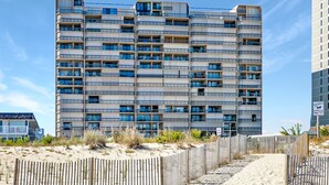 Un edificio de varios pisos con numerosos balcones, una playa arenosa en primer plano y un cielo azul despejado.