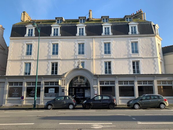 A multi-story building with a classic architectural style, featuring a central entrance with a decorative archway. There are several windows on each floor, and the building has a flat roof with some greenery on top. - Comfortable Apartment (ST MALO)