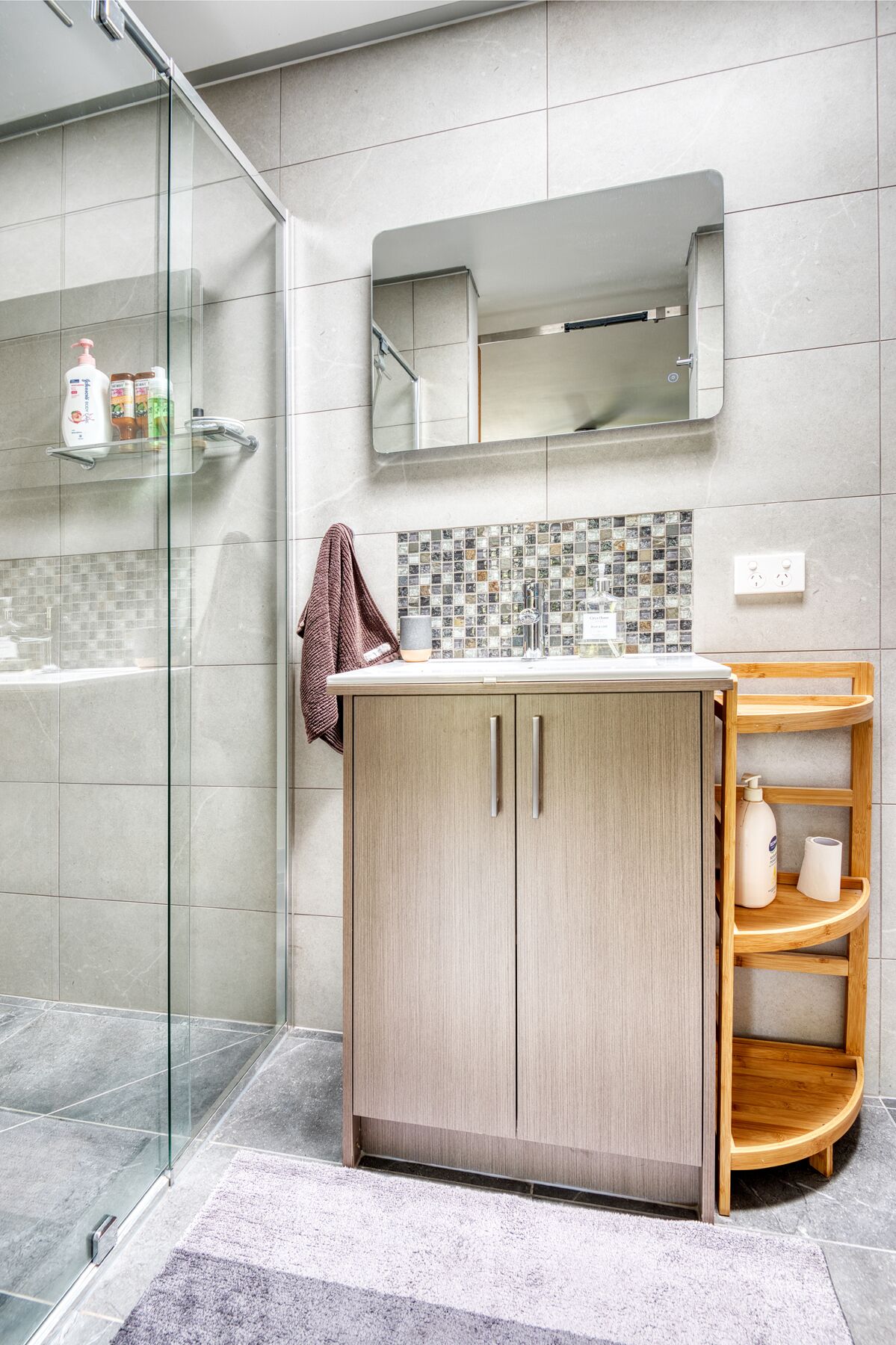 Un baño moderno con cabina de ducha de vidrio, tocador de madera y salpicadero de azulejos, además de una estantería de madera.