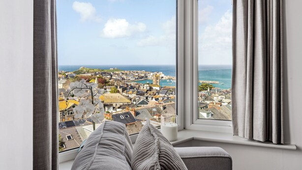 A room with a large window overlooking a coastal town, a bed with pillows, and a glass of milk on the windowsill.