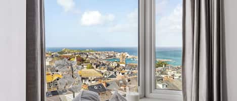 Ein Zimmer mit großem Fenster und Blick auf eine Küstenstadt, einem Bett mit Kissen und einem Glas Milch auf der Fensterbank.