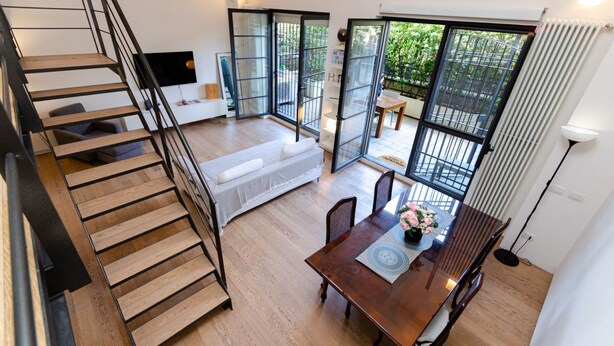 Un interior moderno con una escalera de madera, una zona de comedor con mesa y sillas, y un sofá.