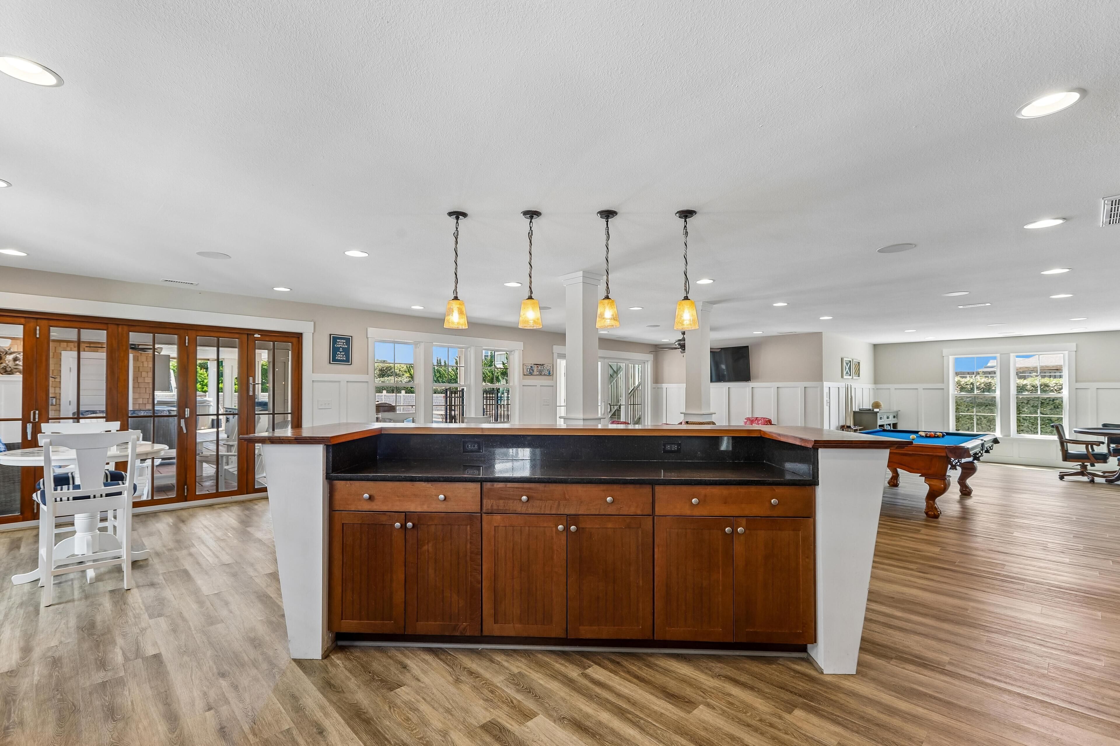A spacious kitchen with wooden cabinets, a central island, pendant lighting, and a pool table in the background.