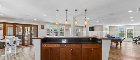 A spacious kitchen with wooden cabinets, a central island, pendant lighting, and a pool table in the background.
