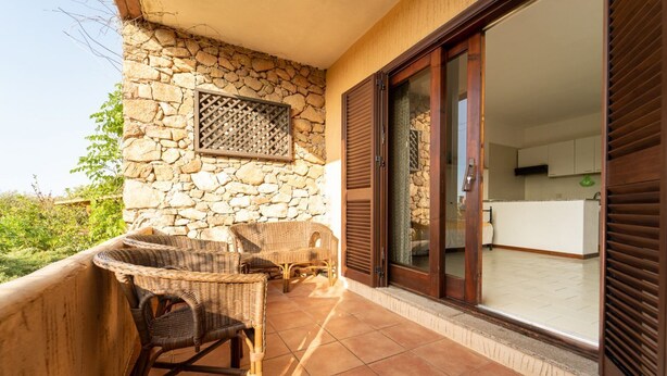 A balcony with wicker furniture, a stone wall, and a sliding glass door leading to a kitchen. - VivimiHome–Mare e relax nel cuore di Porto Rotondo (olbia)