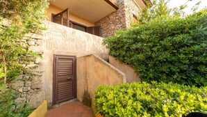 A narrow courtyard with a stone wall, a wooden door, and lush green bushes. - VivimiHome–Mare e relax nel cuore di Porto Rotondo (olbia)