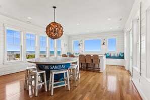 A spacious kitchen with a round table labeled 'THE SANDBAR,' wooden flooring, white cabinetry, and large windows offering ocean views.