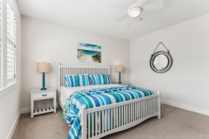 A bedroom with a white crib, a bed with striped bedding, a wall-mounted mirror, and a beach-themed artwork. - Knot Home -- 16BR OF in Corolla with Pool, Elevator, Theater, Game Room and More! (Corolla)