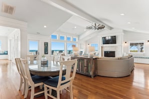 A spacious living room with a large round table, white chairs, a gray sofa, and a fireplace. - Knot Home -- 16BR OF in Corolla with Pool, Elevator, Theater, Game Room and More! (Corolla)