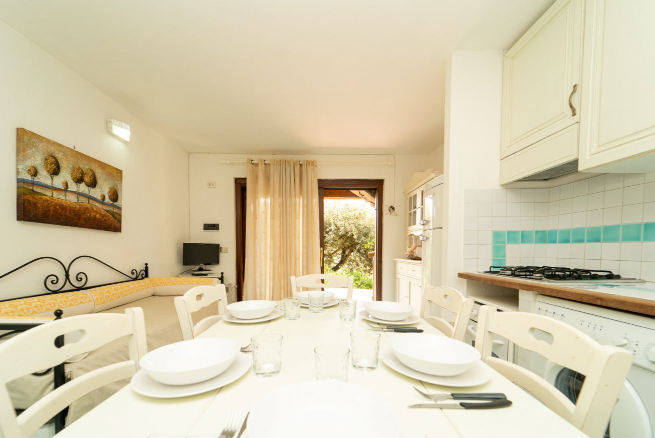 Un comedor con una mesa dispuesta para cuatro personas, sillas blancas, un cuadro en la pared y una cocina con armarios blancos y un salpicadero de azulejos.