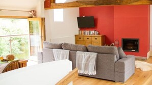 Ein gemütliches Wohnzimmer mit grauem Sofa, Holzbalken, einer roten Akzentwand, einem Kamin und einem Fernseher.