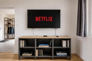 A flat-screen TV mounted on a wall with the word 'NETFLIX' displayed in red letters. Below the TV, there is a wooden console with shelves containing books and a remote control.