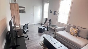 A modern living room with a grey sofa, a black ottoman, a desk with a computer, and a chair. There are two windows with blinds, a ceiling light fixture, and a cityscape picture on the wall.