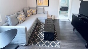 A modern living room with a grey sofa, a black ottoman, a black TV stand, and a framed poster on the wall.