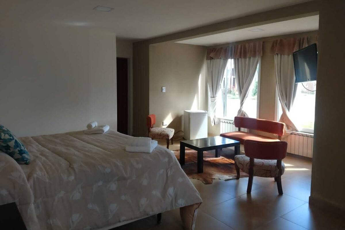 A hotel room with a bed, a small table, a chair, and a window with curtains.