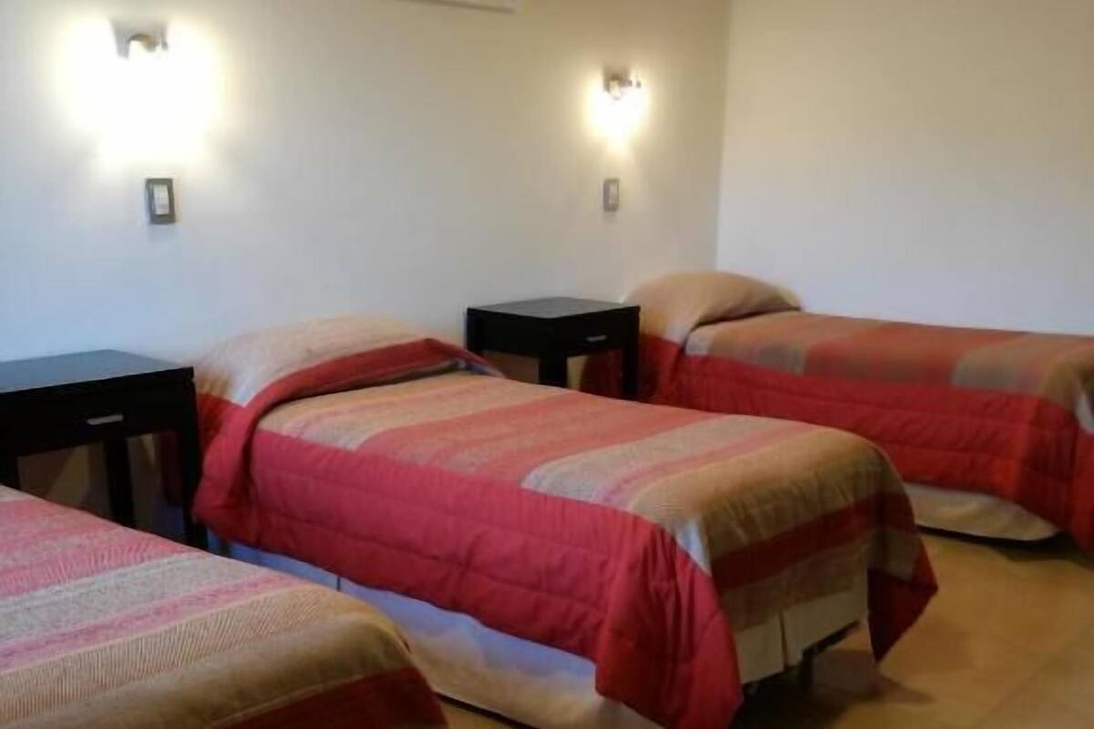 A hotel room with three beds, each with a red and beige bedspread. There are two wall-mounted lights and a framed picture on the wall.