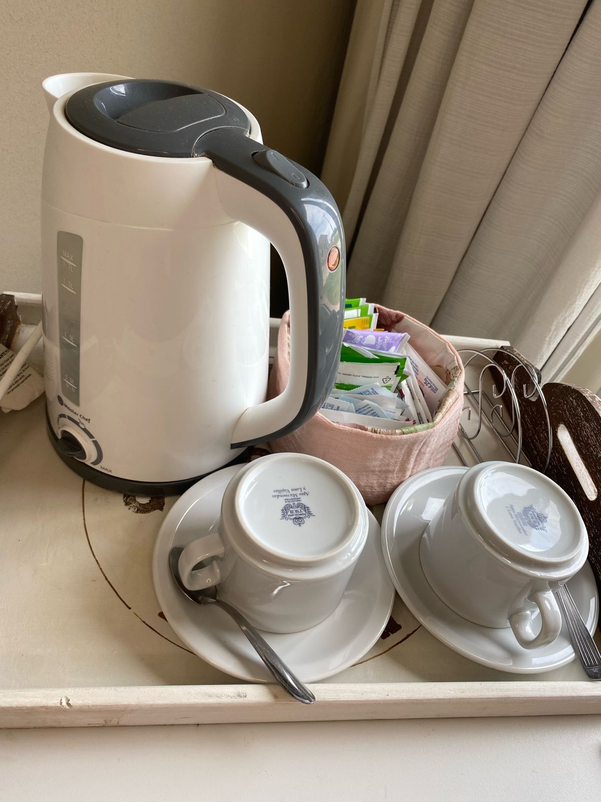 A white kettle, two white cups with saucers, and a pink bag containing various items on a tray.