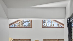 Ein geräumiges Zimmer mit Holzboden, einem großen Bett mit flauschiger weißer Decke, einem Holztisch mit Stühlen und großen Fenstern mit Blick auf Bäume.