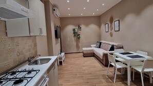 A modern kitchen with a gas stove, a sink, and a small countertop. There is a dining area with a table and chairs, and a sofa in the background. - PICCOLE DONNE (Naples)