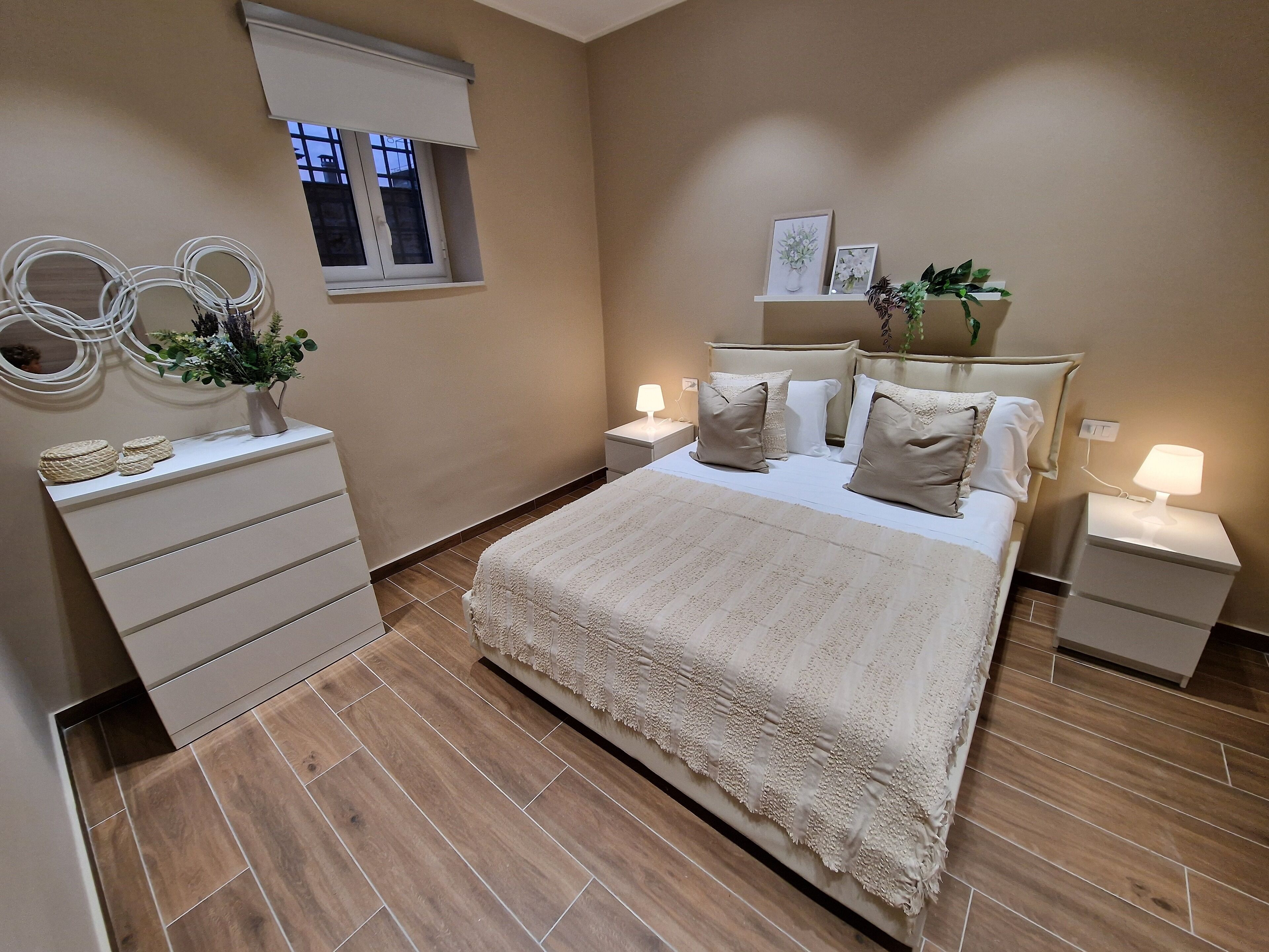 A bedroom with a large bed, bedside tables with lamps, a dresser, a window, and wall decor.