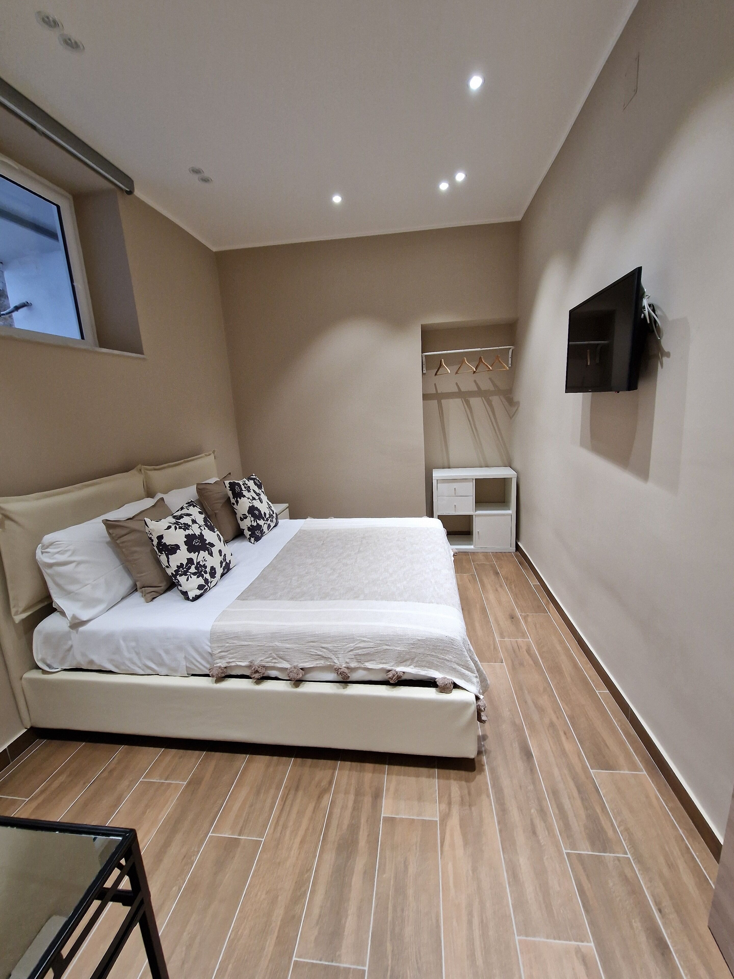 Un dormitorio con una cama, un televisor montado en la pared, una mesita de noche y una ventana.