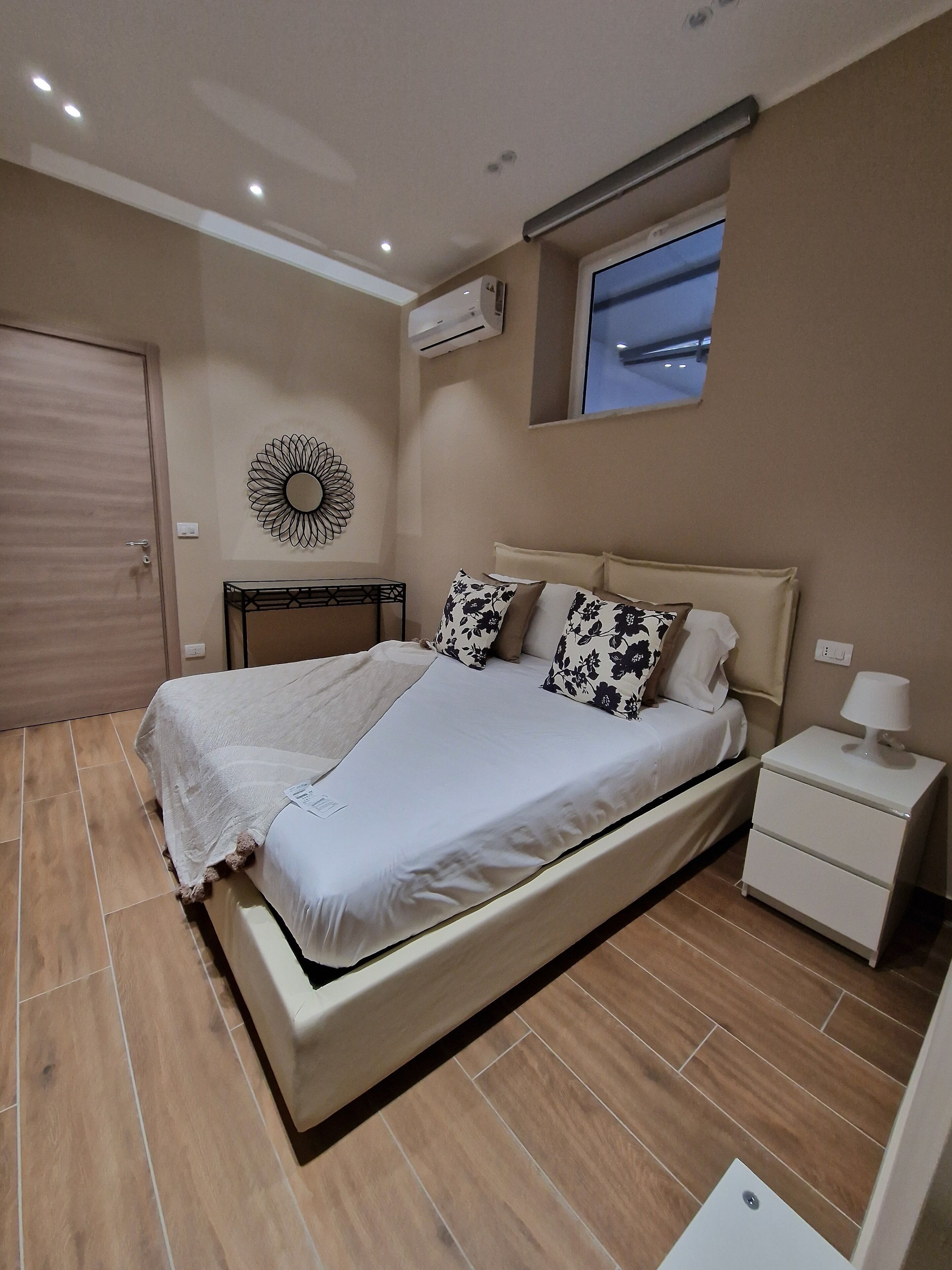 A bedroom with a bed, bedside table, nightstand, wall-mounted air conditioner, and a decorative wall piece.