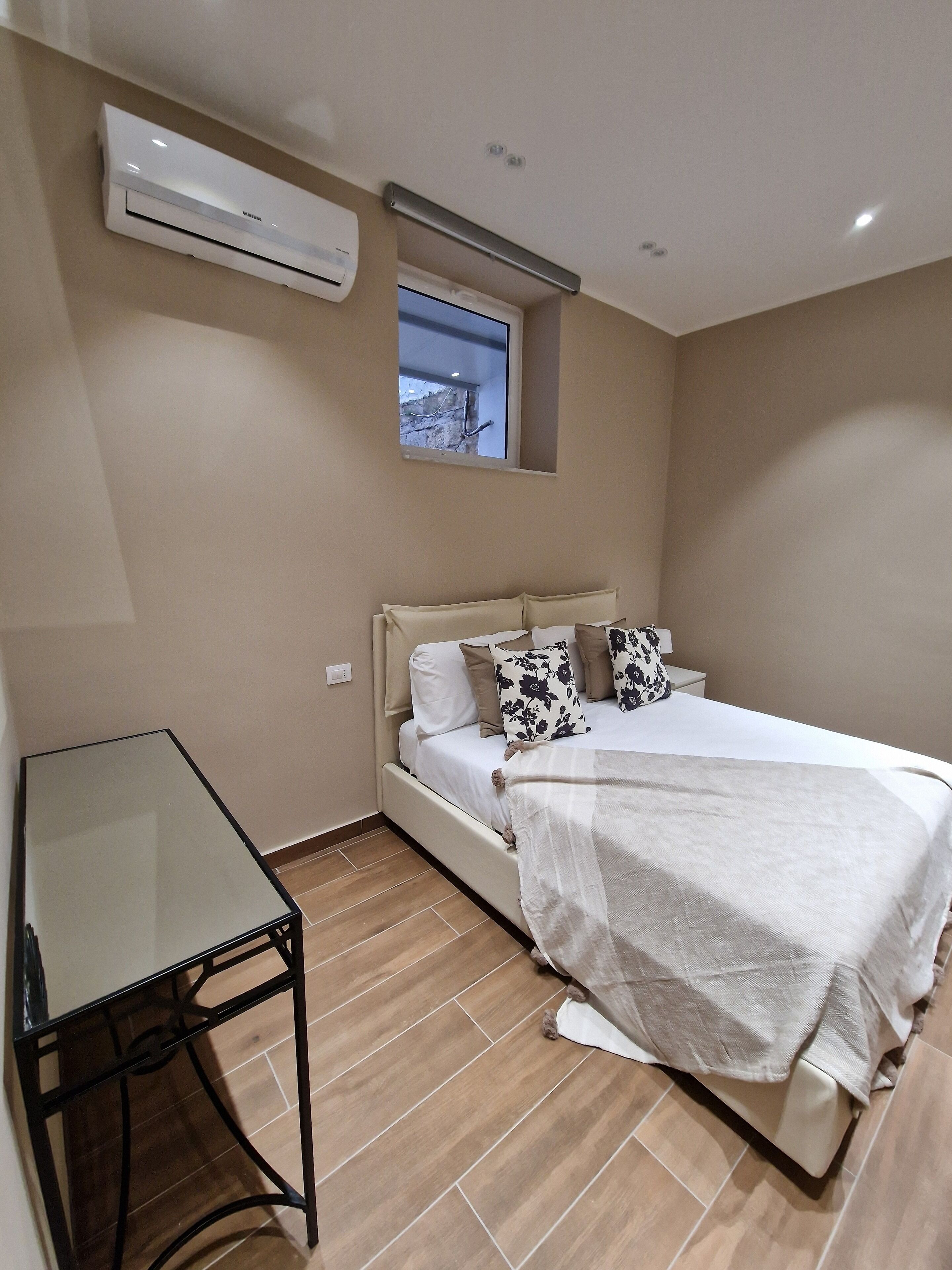 A bedroom with a bed, bedside table, air conditioning unit, window, and a view of the outdoors.