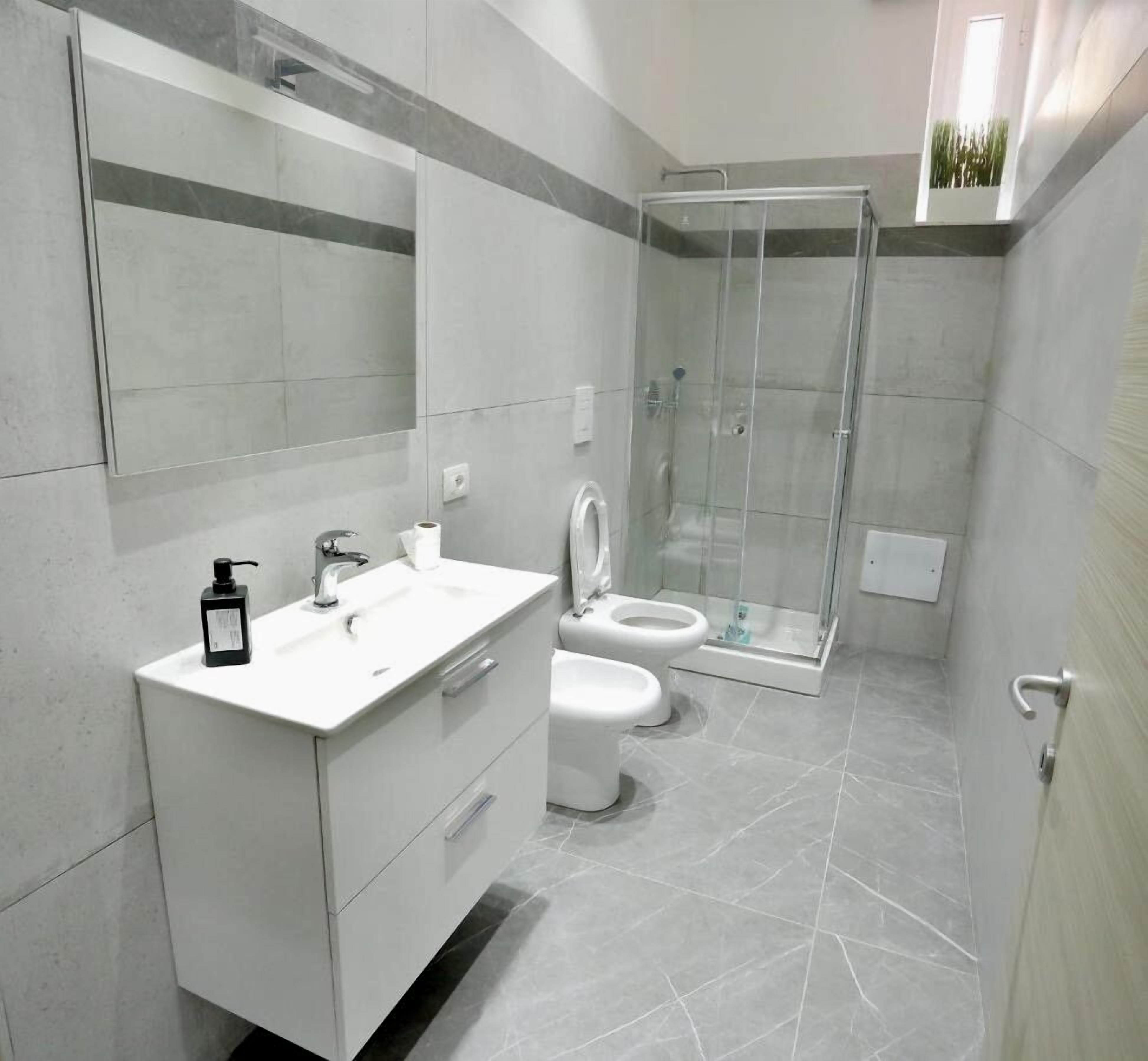 A modern bathroom with a white vanity, two toilets, a glass shower enclosure, and a large mirror.