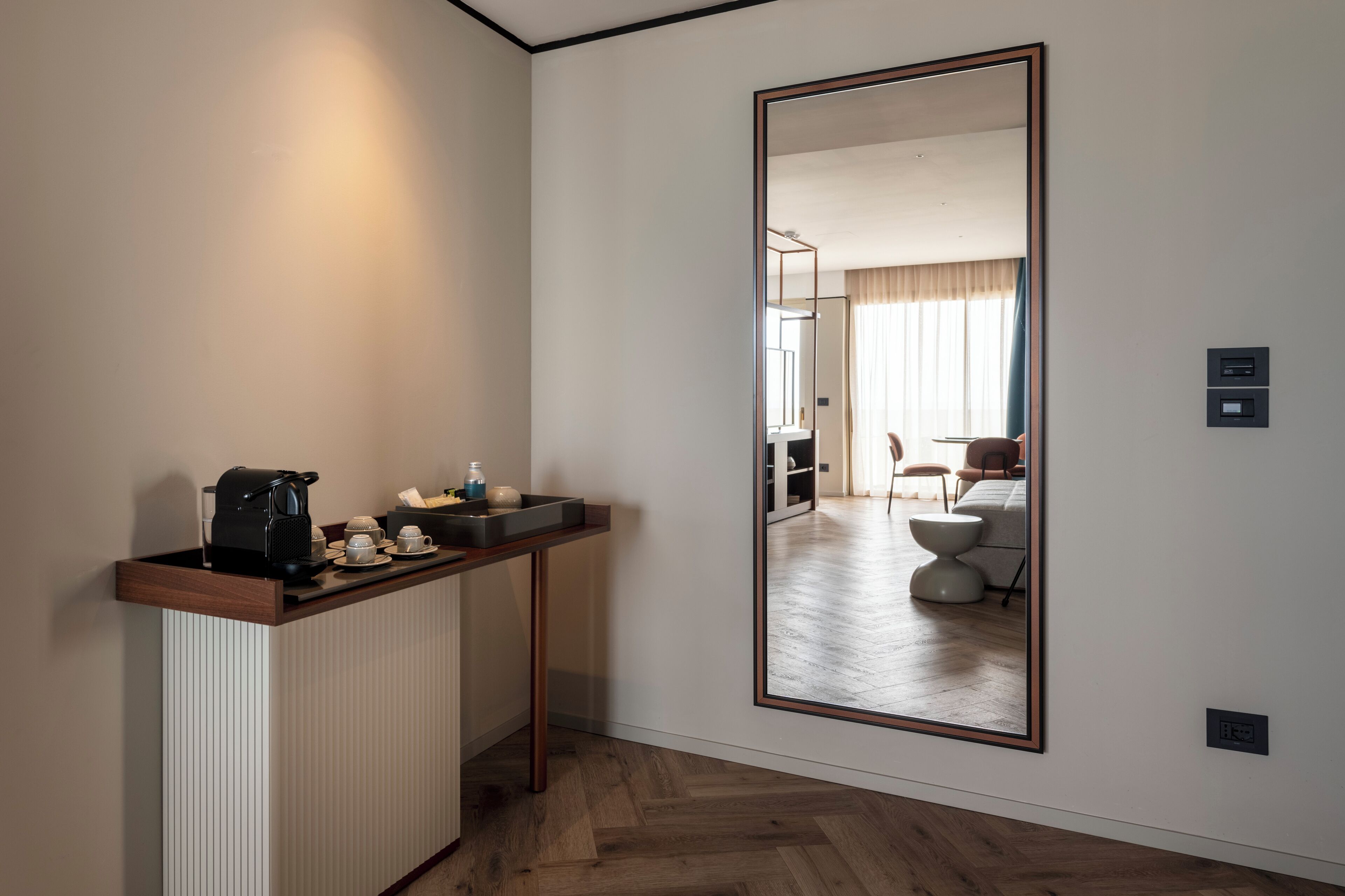 A hotel room with a wooden floor, a small kitchenette with a coffee machine, and a large mirror reflecting the room.