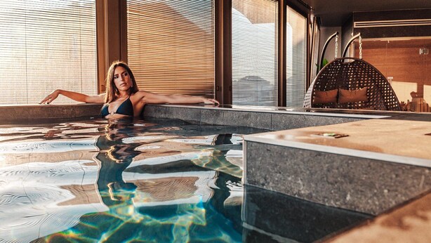 Una persona relajándose en una piscina interior moderna con agua cristalina, borde de piedra y zona de terraza de madera.
