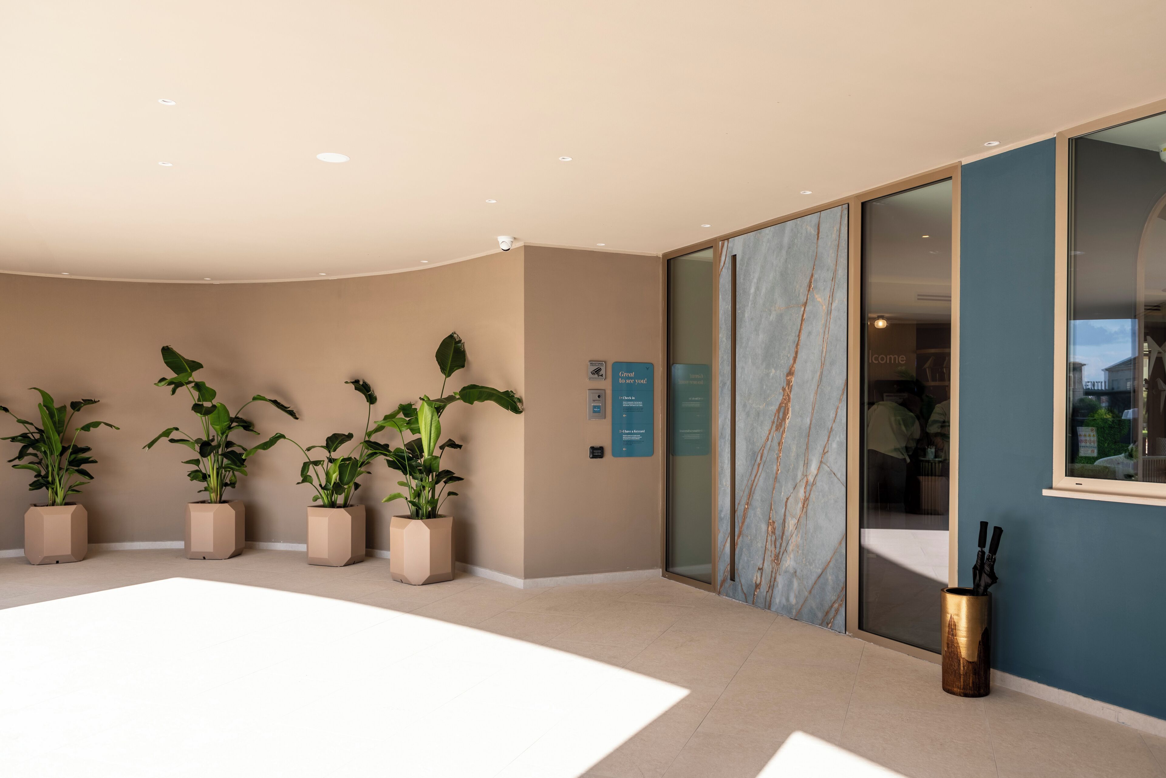A modern hotel lobby with a curved ceiling, beige walls, and a large window. There are three potted plants and a trash can.
