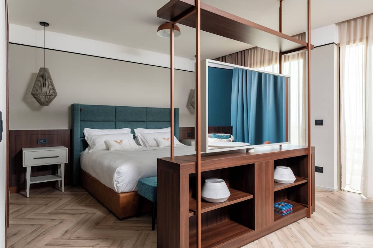 Une chambre d’hôtel moderne avec un grand lit, une table de chevet en bois, un miroir et une étagère intégrée agrémentée d’objets décoratifs.