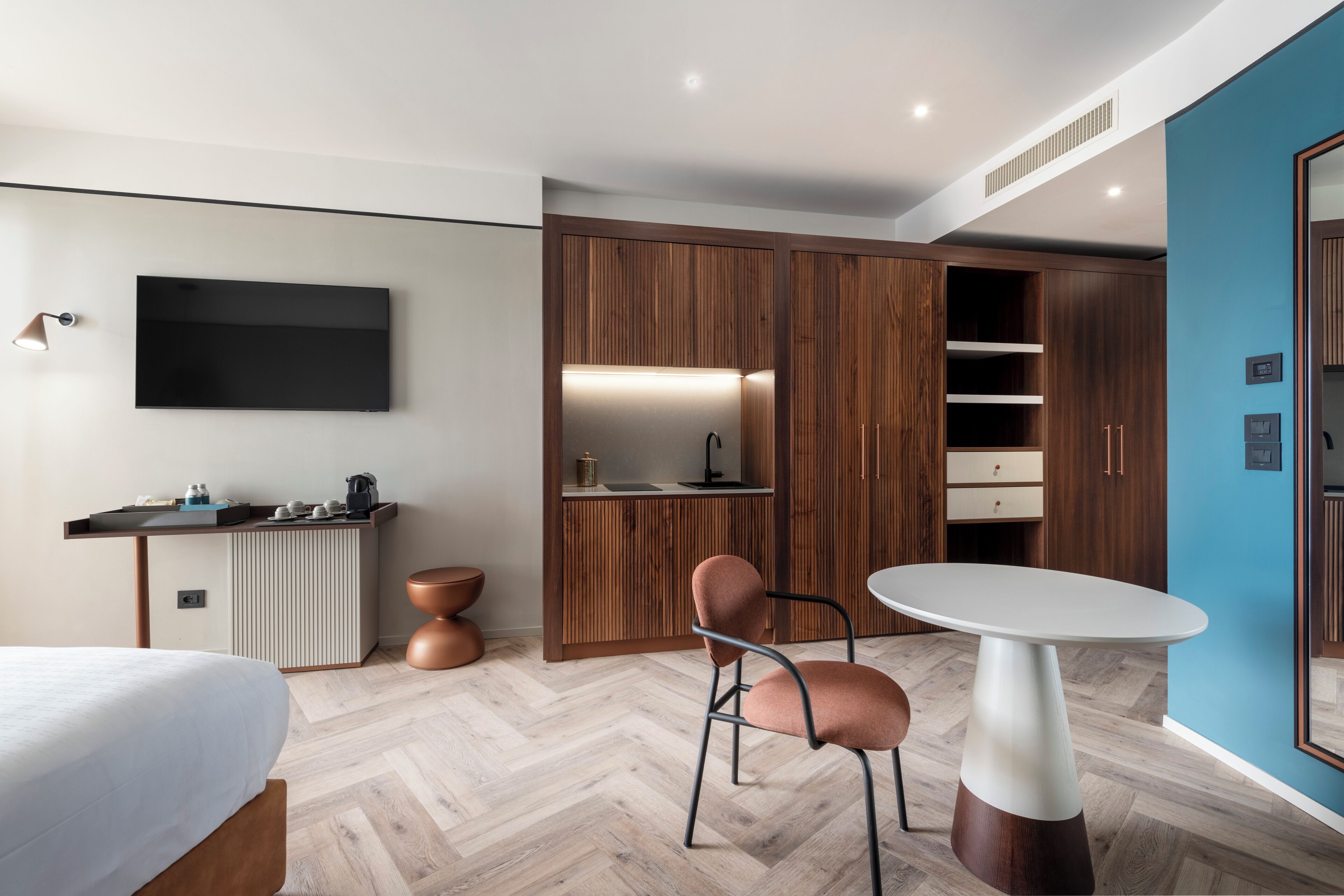 A modern hotel room with a wooden wardrobe, a flat-screen TV, a dining table, and a bed.