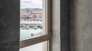 Un baño con un ventanal grande, ducha y vista a un puerto con barcos.