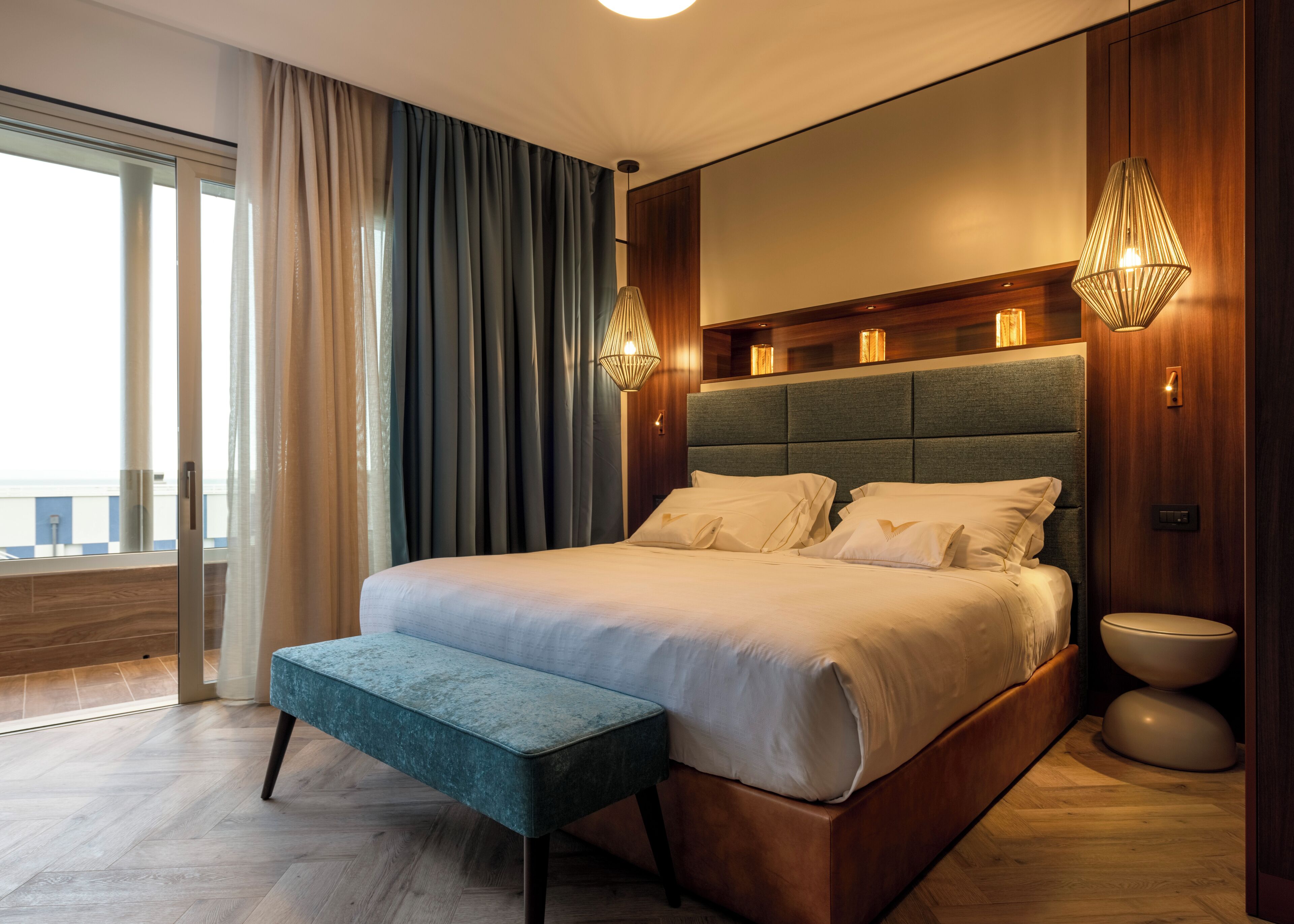 A modern bedroom with a large bed, a bench, a view of the city, and a wooden headboard.